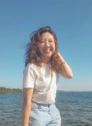 brown-haired woman smiling toothily at the camera with one hand up, wearing a purple cardigan and white T-shirt. in the background are cherry blossom trees