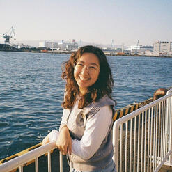 brown-haired woman smiling toothily at the camera with one hand up, wearing a purple cardigan and white T-shirt. in the background are cherry blossom trees